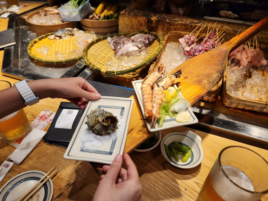 점원이 긴 나무 주걱으로 손님에게 뿔소라 구이를 전달하는 모습.