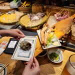 점원이 긴 나무 주걱으로 손님에게 뿔소라 구이를 전달하는 모습.