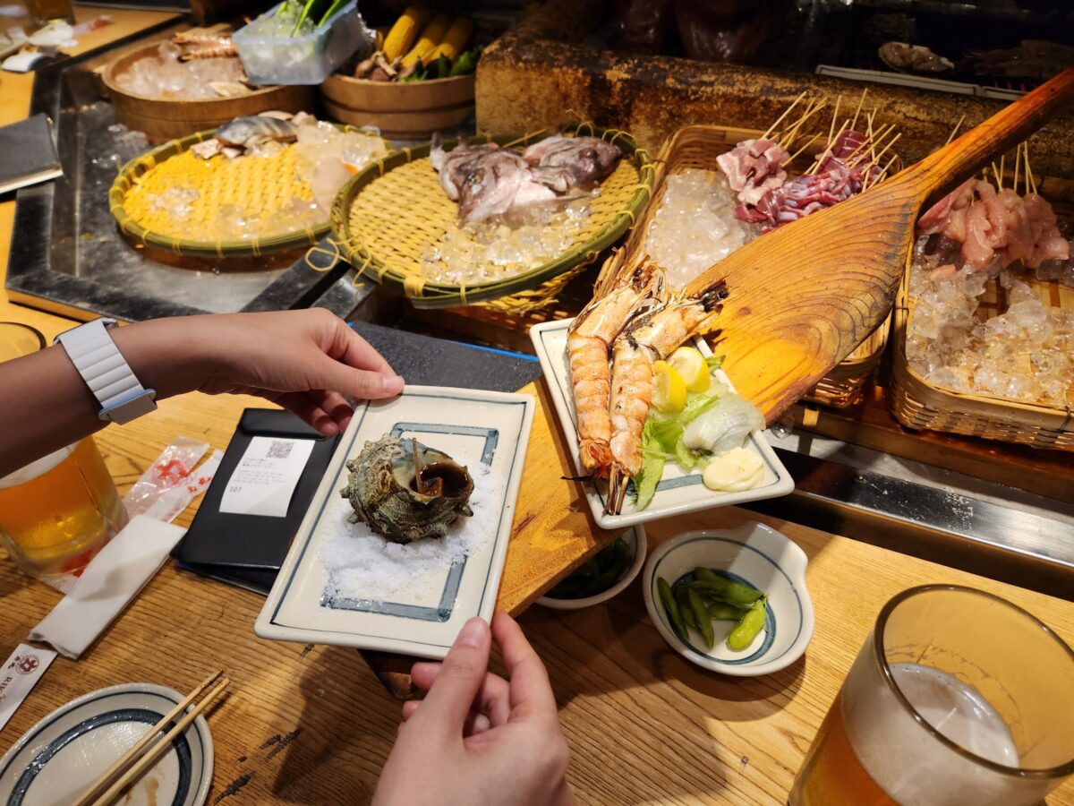 점원이 긴 나무 주걱으로 손님에게 뿔소라 구이를 전달하는 모습.