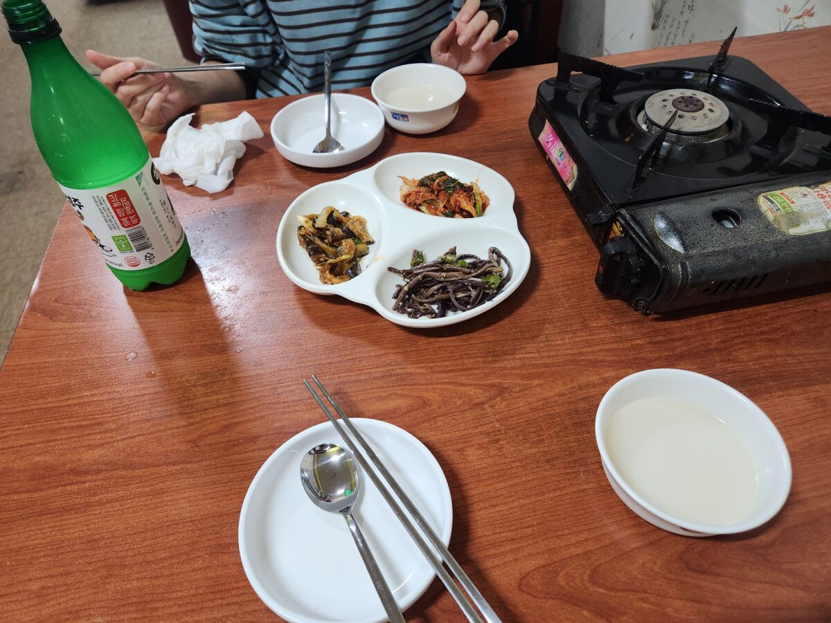 식탁 위에 차려진 막걸리병, 기본 반찬 3종과 빈 막걸리 잔이 놓인 모습.