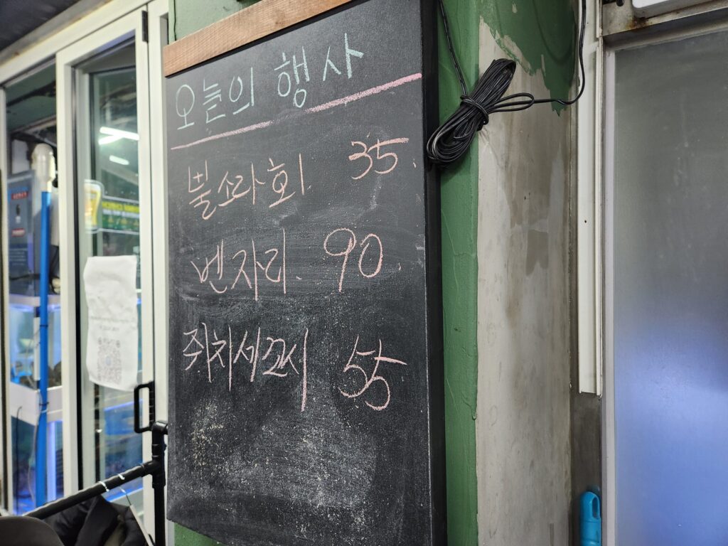 칠판에 '오늘의 행사'와 함께 볼락회, 벤자리, 쥐치세꼬시 메뉴와 가격이 적혀 있는 모습.