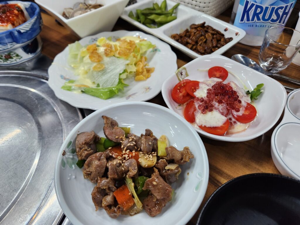 테이블 위에 차려진 토마토 치즈 샐러드, 닭똥집 볶음, 콘푸레이크 샐러드, 번데기 등 다양한 밑반찬.