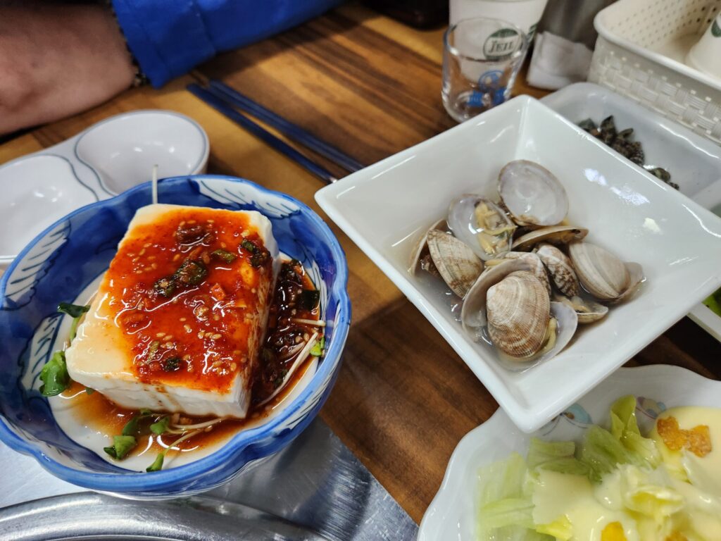 양념장이 올라간 순두부와 접시에 담긴 조개 요리 등 기본 안주 구성.