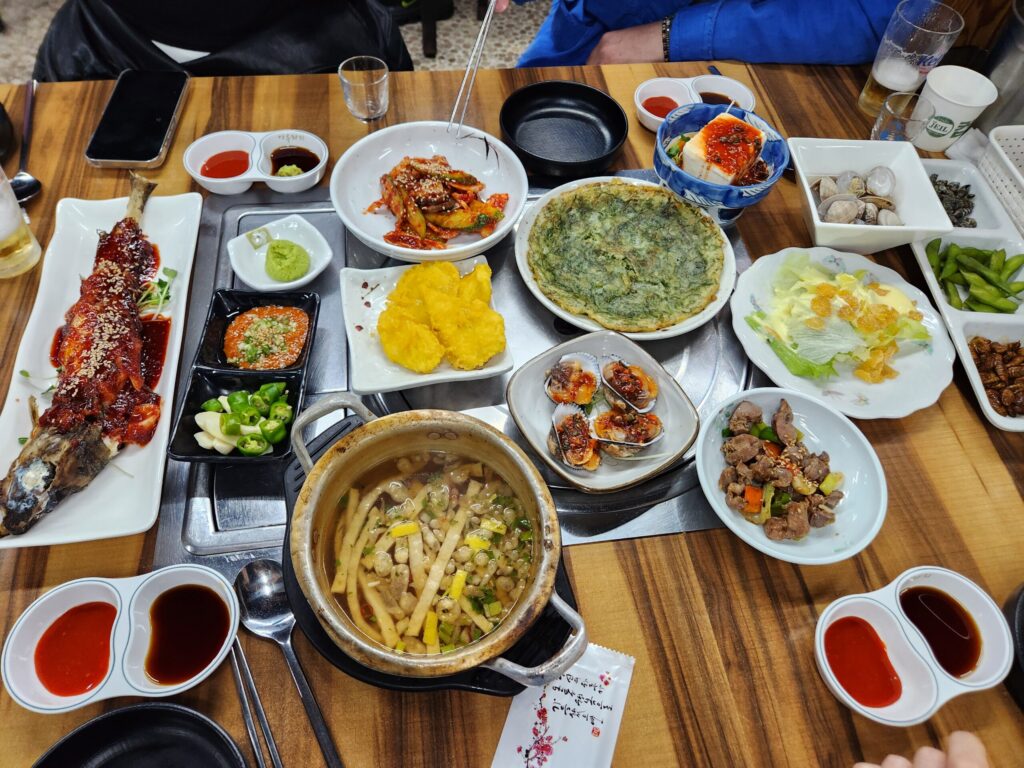 테이블 위에 코다리찜, 파래전, 피꼬막, 유린기 등 다양한 기본 반찬들이 가득 차려진 모습.