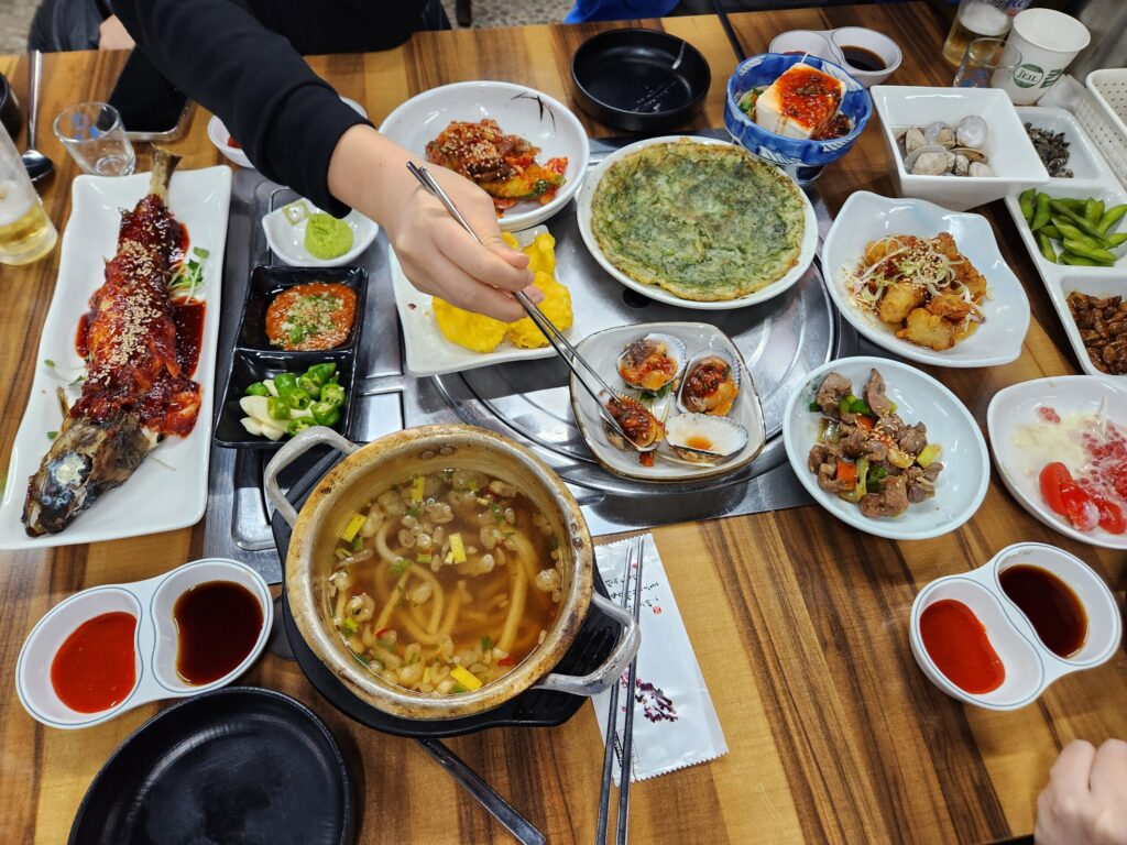 여러 가지 스끼다시가 차려진 테이블에서 사람이 젓가락으로 피꼬막을 집고 있는 모습.