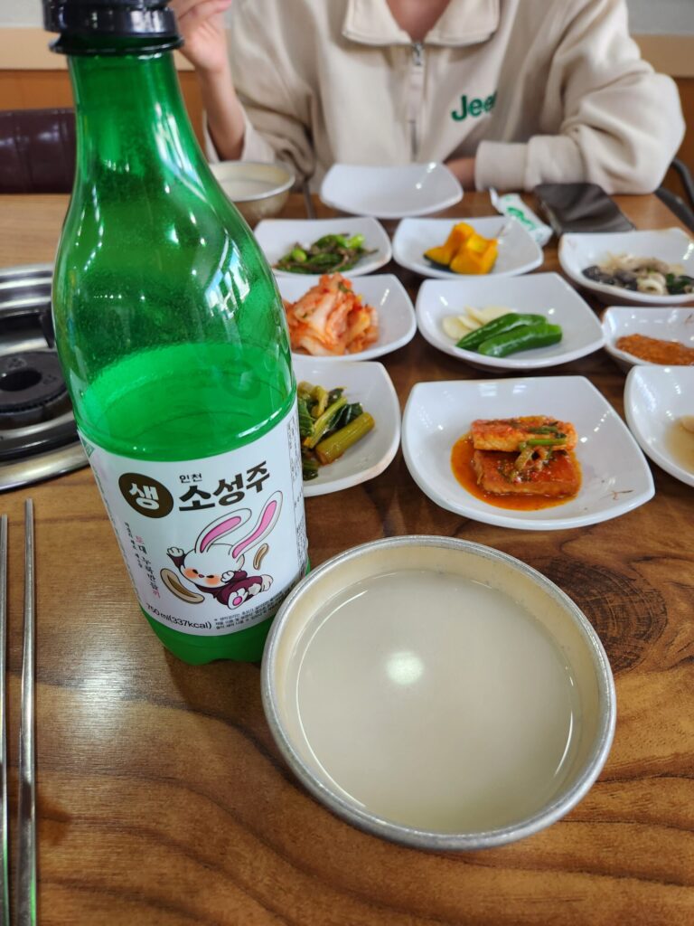 막걸리 병과 따라놓은 잔, 그리고 주변의 밑반찬들이 있는 테이블.