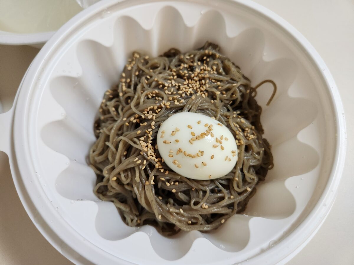 포장 용기에 담긴 메밀면과 삶은 계란, 깨가 뿌려진 물막국수.